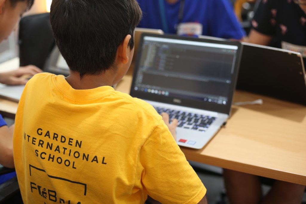 A kid wearing a yellow GIS tshirt coding on a laptop